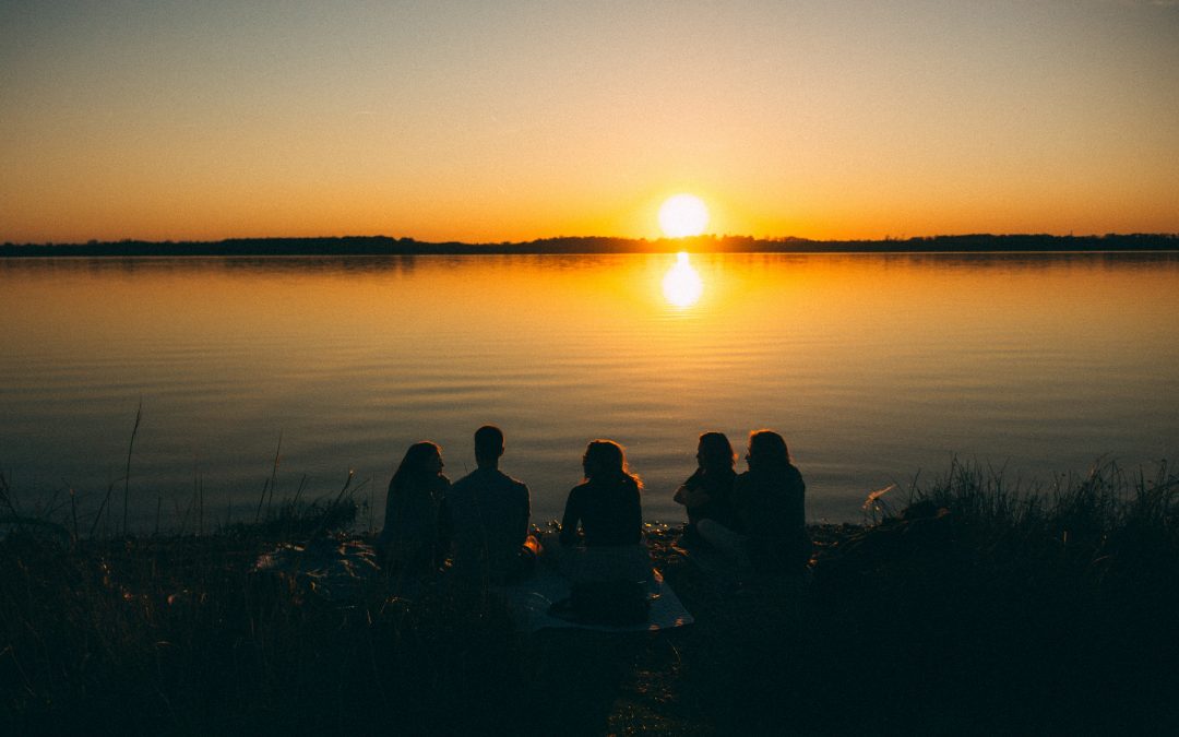 a group of friends gather on the beach with the sun setting on the horizon.