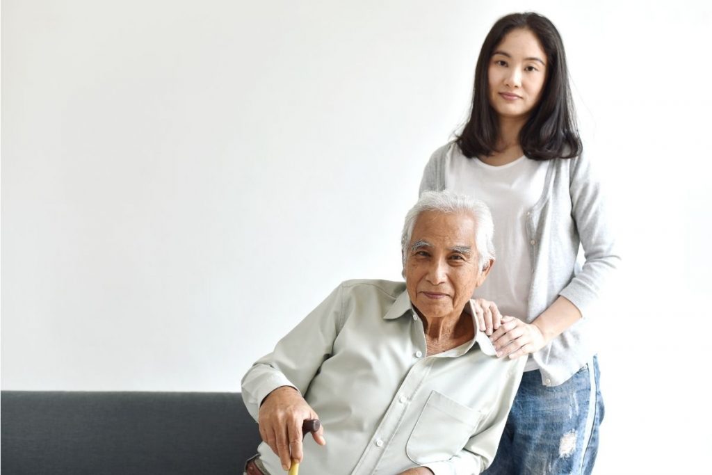 Younger Asian woman standing behind an older father looking figure who is sitting in a chair while holding a cane. Both look content. 