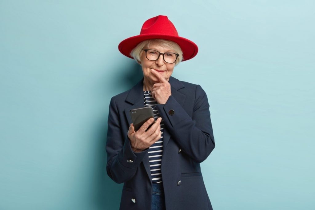 smart looking older adult female wearing fashionable clothing and a red hat holding a smart phone. Practice being assertive to prevent abusive caregiver situations