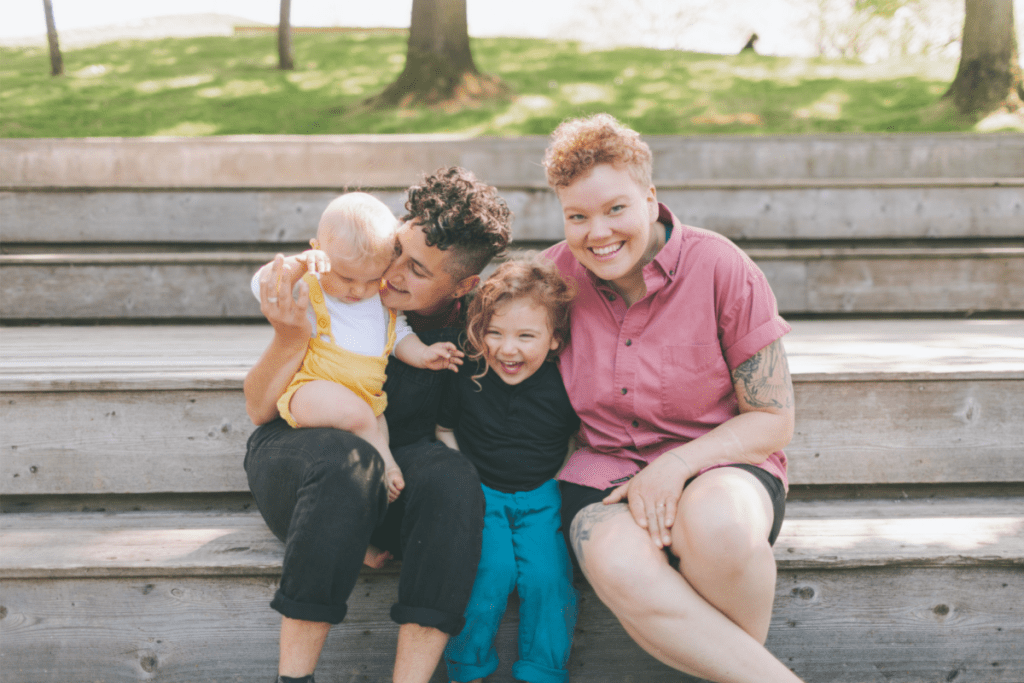 two mothers snuggle their toddler and baby while sitting on a park bench. They are considering making an estate plan for their blended family
